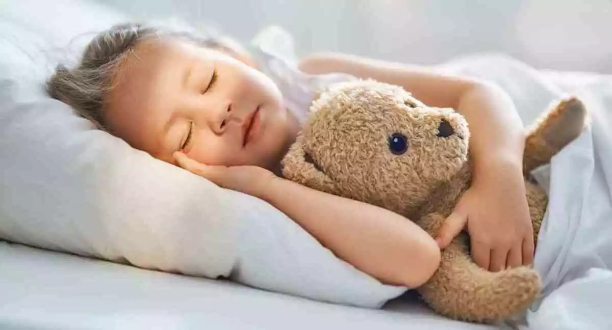Adorable little child is sleeping in the bed with her toy. The girl is hugging the teddy bear.