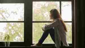 Thoughtful girl sitting on sill embracing knees looking at window